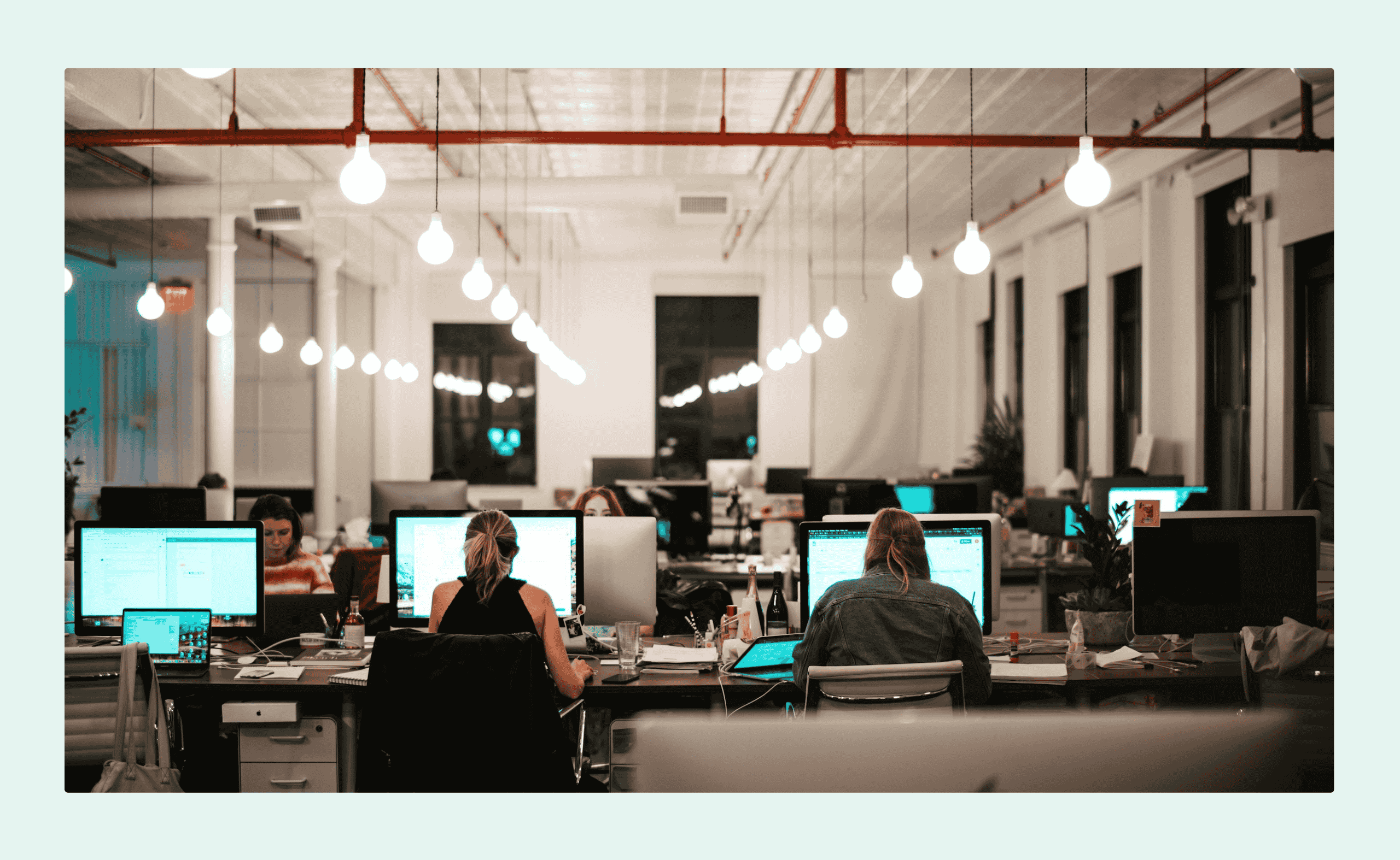 80+ Hybrid Work Statistics in 2026: Productivity & Preferences Open-plan office with people working at shared desks, surrounded by monitors and warm hanging lights in a modern coworking space.