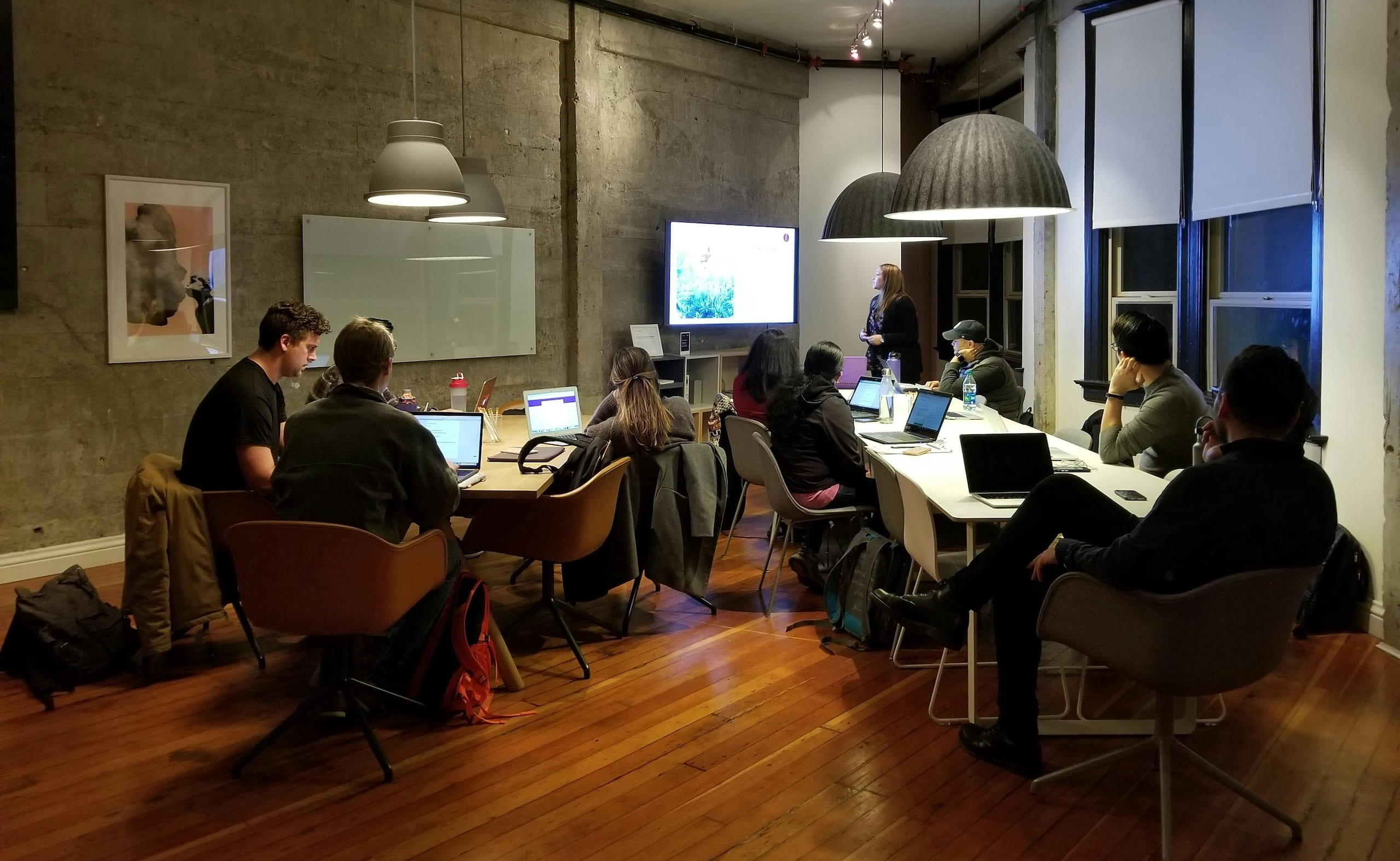 People sitting around tables with laptops during a presentation in a modern, industrial-style meeting room.