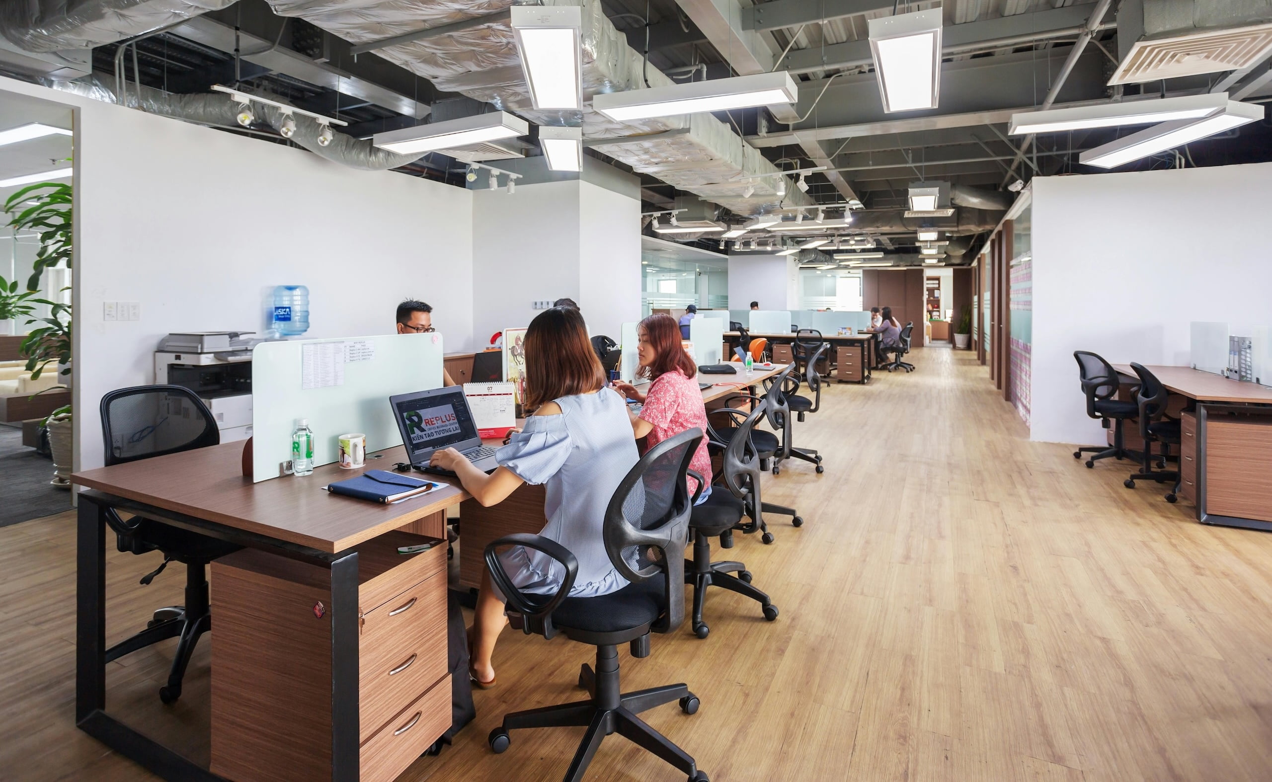 Open-plan office with employees working at rows of desks, featuring modern lighting and a clean, spacious layout.
